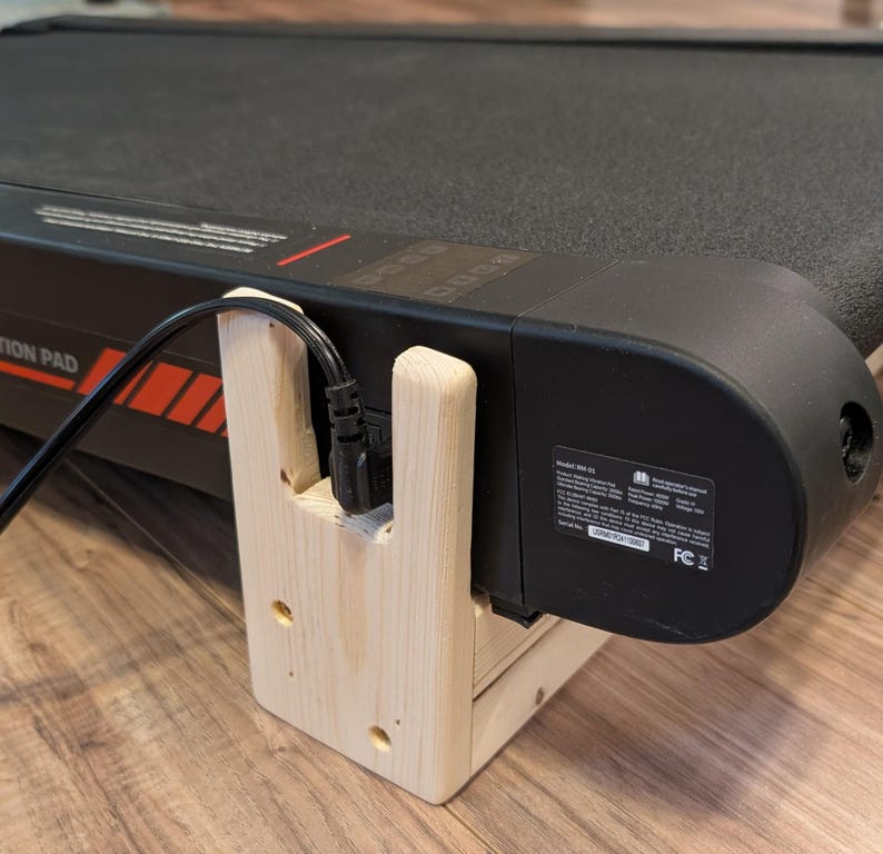 May include: Close-up of a black treadmill with a wooden support structure. The treadmill has a black belt and a digital display. The wooden support is light-colored and holds the power cord.