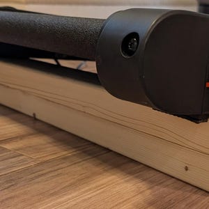 May include: A black and gray exercise machine with a textured roller is mounted on a light-colored wooden base. The base is made of unfinished wood and is rectangular. The machine has a black plastic casing.