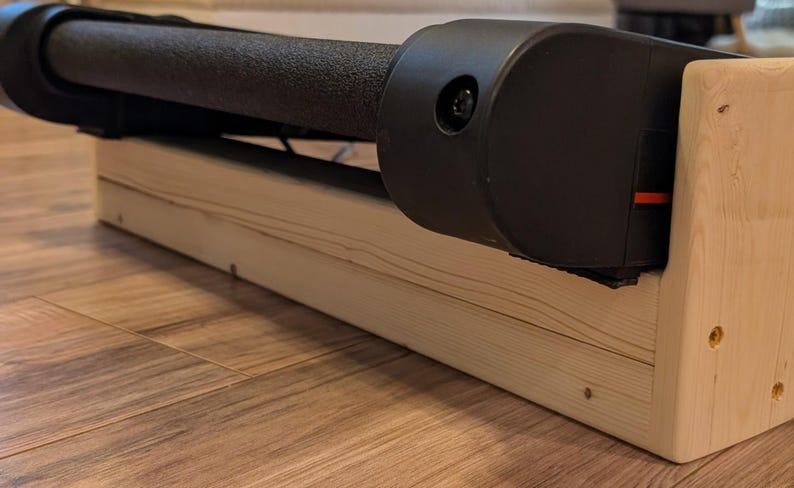 May include: A black and gray exercise machine with a textured roller is mounted on a light-colored wooden base. The base is made of unfinished wood and is rectangular. The machine has a black plastic casing.
