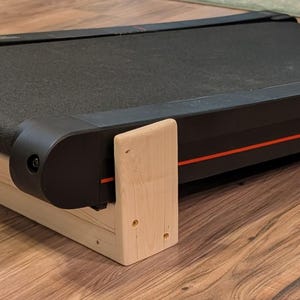 May include: A black treadmill with a red stripe and text on the side, resting on a light-colored wooden platform. The treadmill belt is black and the frame is black with red accents. The wooden platform is made of light-colored wood.