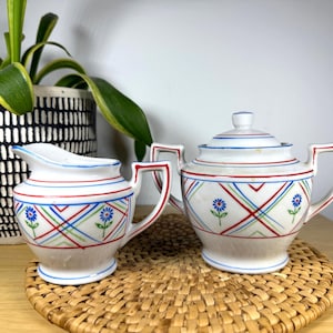 May include: A white ceramic creamer and sugar set with a blue, red, and green geometric pattern and blue flower accents.