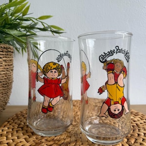 May include: Two clear glass tumblers with Cabbage Patch Kids illustrations. One shows a girl in a red dress skipping, the other shows a girl upside down. The Cabbage Patch Kids logo is printed on each glass.