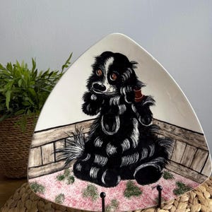 May include: A triangular ceramic plate with a black and white illustration of a dog sitting on a pink and green patterned rug. The dog has brown eyes and is wearing a red collar. The plate is on a stand.