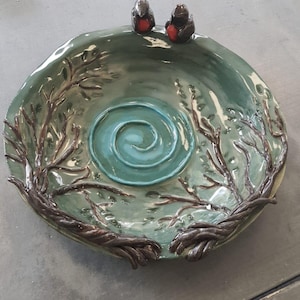 Puede incluir: Un bebedero para pájaros de cerámica artesanal con un esmalte verde azulado. El cuenco presenta un diseño de espiral central y está adornado con ramas esculpidas y dos pequeñas figuras de pájaros con pechos rojos. El diseño evoca un tema natural y de bosque.