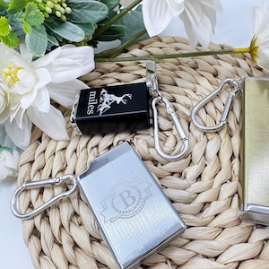 Op de afbeelding: Een verzameling metalen sleutelhangers, waaronder een zwarte rechthoekige met het woord "miles" en een berg- en hertenafbeelding. Een andere sleutelhanger is zilverkleurig met de letter "B" in een lauwerkrans. Alle sleutelhangers hebben zilveren karabijnhaakclips.