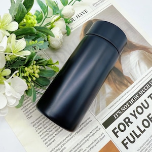 May include: A sleek, black insulated water bottle with a matte finish. The cylindrical bottle has a matching black lid and appears to be made of stainless steel. The bottle is shown on a newspaper background with white flowers.