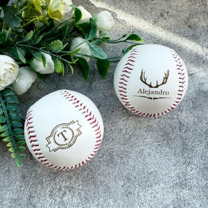 Könnte beinhalten: Zwei weiße Baseballs mit roten Nähten. Ein Baseball hat ein goldenes Hirschkopfdesign mit dem Namen "Alejandro" darunter gedruckt. Der andere Baseball hat einen goldenen Kranz mit dem Buchstaben "T" darin.