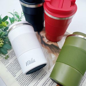 May include: Four insulated travel mugs in black, red, white, and olive green. Each mug has a stainless steel rim and a textured silicone band for grip. The white mug has the name "Mila" printed on it.