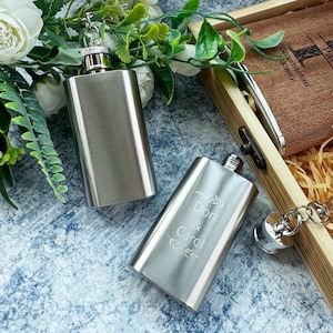 Op de afbeelding: Twee zilveren hipflasks met gegraveerde tekst. Een flesje zegt "Don & Carol" met een bloemmotief. De andere fles is effen. Beide flesjes hebben een zilveren dop en een zilveren ketting.