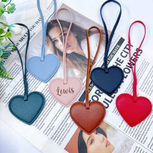 May include: A collection of heart-shaped leather tags in various colors: teal, light blue, pink with "Lewis" printed on it, brown, navy, and red. Each tag has a matching leather strap for hanging.