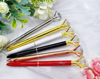 May include: An assortment of diamond-topped pens in silver, gold, white, black, rose gold, and red. The pens have a diamond-shaped crystal at the top and are arranged on a white surface with floral accents. Some pens have text.