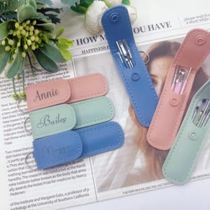 May include: A collection of personalized cosmetic tool sets in pastel colors. Each set includes tools in a matching case, with names like "Annie," "Bailey," and "Maggie" engraved on the tags. The sets are displayed on a newspaper background.