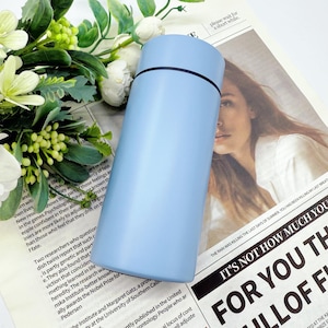 May include: A light blue insulated water bottle with a black band near the top. The cylindrical bottle is made of metal and sits on a newspaper with text and a woman's face. Green and white flowers are in the background.