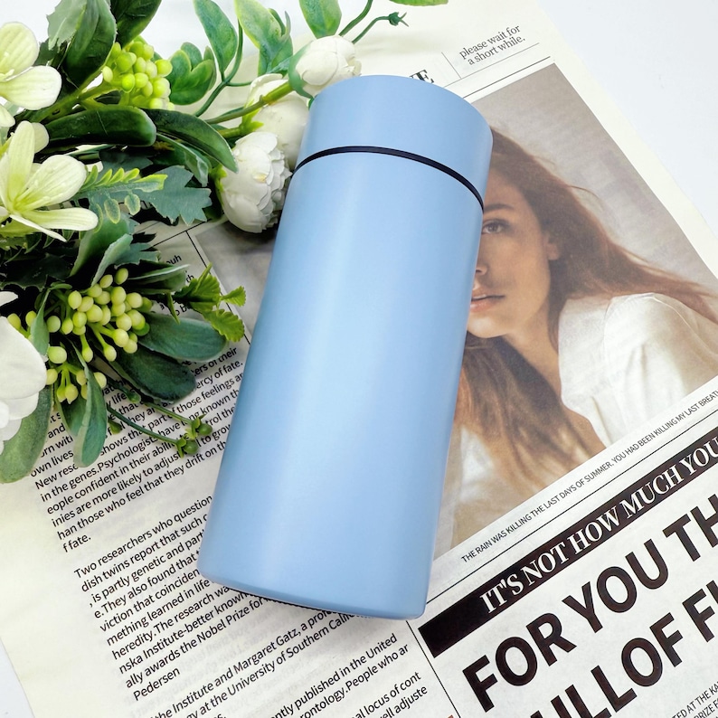 May include: A light blue insulated water bottle with a black band near the top. The cylindrical bottle is made of metal and sits on a newspaper with text and a woman's face. Green and white flowers are in the background.