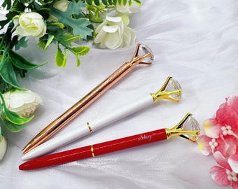 May include: Three diamond-topped pens in rose gold, white, and red. The pens have a diamond-shaped crystal at the top and gold accents. The rose gold pen has the word "Perfect" on it, and the red pen has the word "Sky" on it.