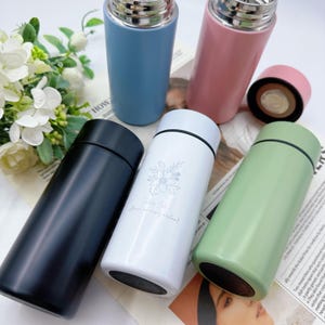 Puede incluir: Una variedad de botellas de agua aisladas de acero inoxidable en varios colores: negro, blanco, azul, rosa y verde. La botella blanca tiene un diseño floral y el texto "Taking Out Almost an empty vessel!"