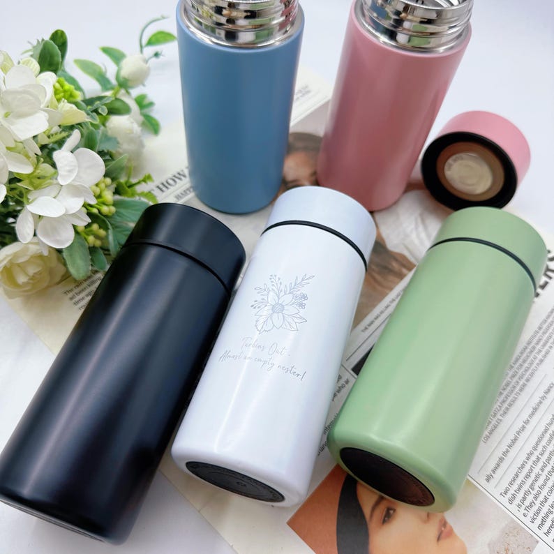 May include: An assortment of insulated stainless steel water bottles in various colors: black, white, blue, pink, and green. The white bottle has a floral design and the text "Taking Out Almost an empty vessel!"