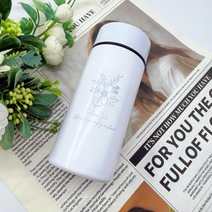 May include: White insulated water bottle with a floral design and the text "Turning Out - Almost an empty nester!". The bottle has a white lid and a black band. The bottle is on a newspaper.
