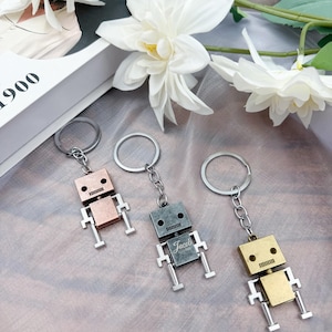 May include: Three robot-shaped keychains in copper, gray, and gold tones. Each robot has a square head with two eye holes and a mouth, and a rectangular body. The gray robot has the name "Jacob" engraved on its body.