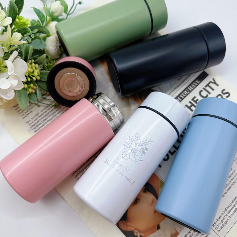 May include: An assortment of insulated stainless steel water bottles in various colors: pink, white, blue, green, and black. The white bottle has a floral design and the text "Working Out... Always an empty water!"