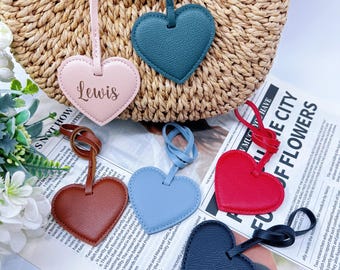 May include: A collection of heart-shaped leather bag charms in various colors, including pink, teal, red, blue, brown, and navy. One charm is engraved with the name "Lewis". The charms are displayed against a woven background.