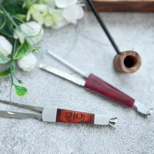 Puede incluir: Dos herramientas de fumar de metal con mangos de madera. La herramienta más grande tiene un mango rojo con el texto "J.O." grabado en él. La herramienta más pequeña tiene un mango marrón con el texto "J.O." grabado en él.