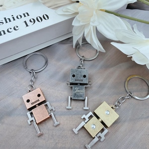 May include: Three robot-shaped keychains in bronze, silver, and gold tones. Each robot has a square head and body with rectangular arms and legs. The silver robot has the name "Jacob" engraved on its body. The keychains are on a silver ring.