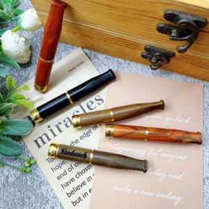 Può includere: Un assortimento di pipe in legno in varie tonalità di marrone e nero, rifinite con fasce color oro. Una pipa è incisa con il nome "Frederick". Le pipe sono disposte su carta colorata chiara con testo motivazionale, vicino a una scatola di legno.