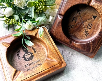 May include: Two wooden square ashtrays with a decorative design. One ashtray has a compass design with the letters R and S. The other ashtray has a silhouette of a man with a top hat and a mustache, with the text "Michael 08.11.2024".