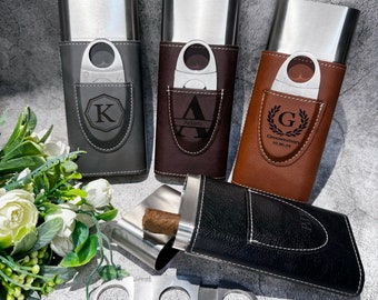 May include: Four personalized cigar cases with a silver finish and leather coverings. The cases have a built-in cutter and a pocket for a cigar. The cases are engraved with initials, names, and dates. The cases are arranged on a gray surface with a bouquet of white flowers.