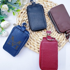Peut inclure: Plusieurs porte-clés en cuir de différentes couleurs : bleu marine, noir, bordeaux et marron. Chaque porte-clés est doté d'une fermeture éclair et d'un anneau métallique. Un porte-clés bleu porte les initiales "M IVI".