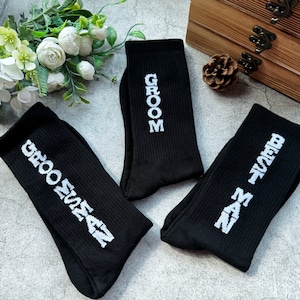 Puede incluir: Tres calcetines negros con texto blanco. Un calcetín dice "Groom", otro dice "Groom Squad" y el último dice "Best Man".