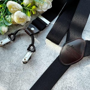 Könnte beinhalten: Schwarze Hosenträger mit einem verstellbaren Lederriemen in Braun und silbernen Metallclips. Der Lederriemen trägt den Text "Manuel Groomsperson 06.18.21" in Prägung.