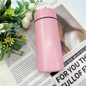 May include: A pink insulated water bottle with a black rim on the lid. The cylindrical bottle is made of stainless steel and is shown on a newspaper background with floral accents.