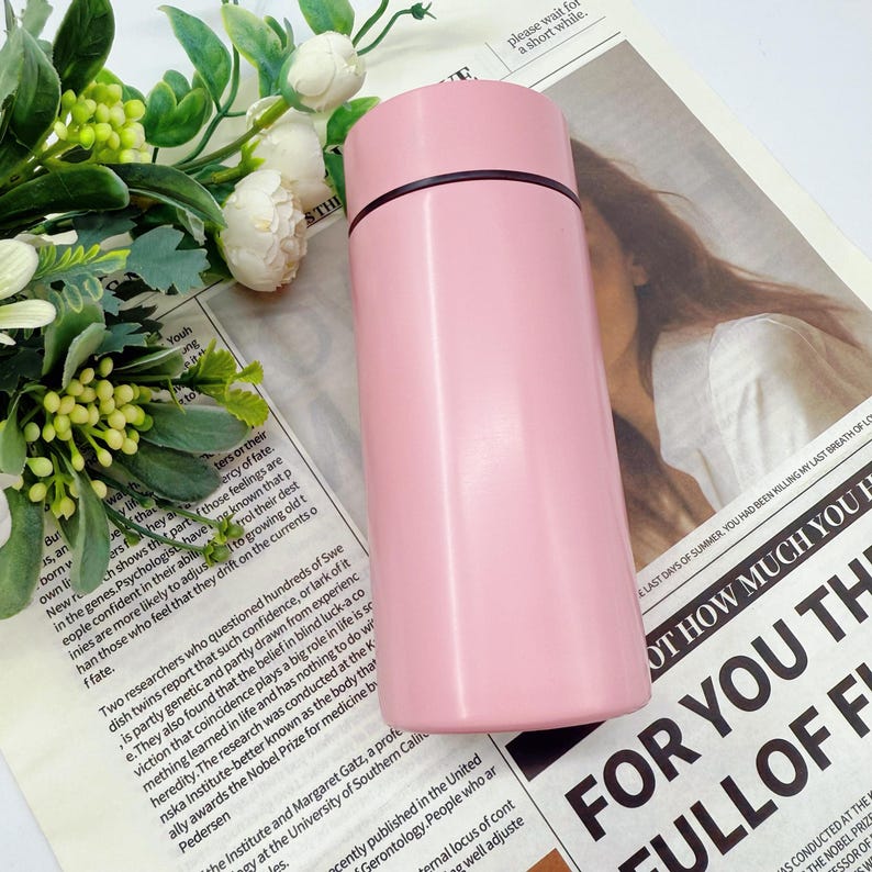 May include: A pink insulated water bottle with a black rim on the lid. The cylindrical bottle is made of stainless steel and is shown on a newspaper background with floral accents.