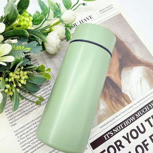 May include: A light green, stainless steel insulated water bottle with a black rim. The bottle is cylindrical with a slightly curved shape. The background includes a newspaper and white flowers.