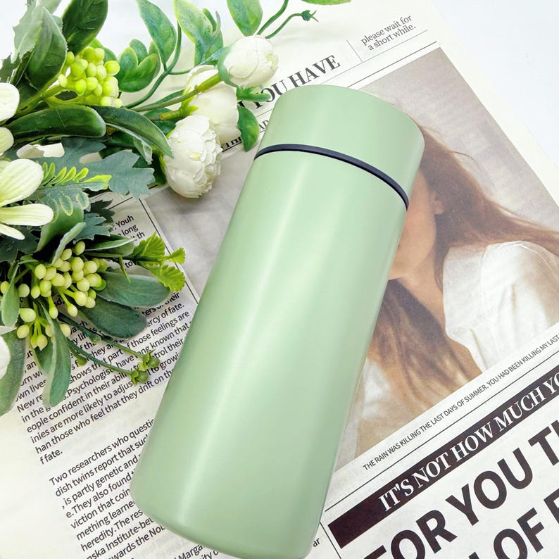 May include: A light green, stainless steel insulated water bottle with a black rim. The bottle is cylindrical with a slightly curved shape. The background includes a newspaper and white flowers.