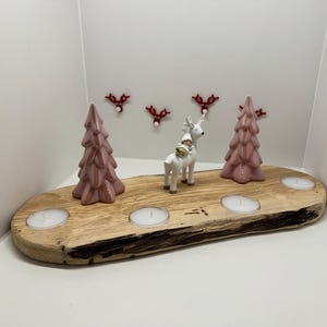 Könnte beinhalten: Ein Kerzenhalter aus Holz mit vier Teelichthaltern und zwei rosa Keramikkristbäumen. Eine weiße Keramik-Rentierfigur befindet sich in der Mitte des Halters. An der Wand hinter dem Halter befinden sich fünf rote Rentierkopf-Ornamente.