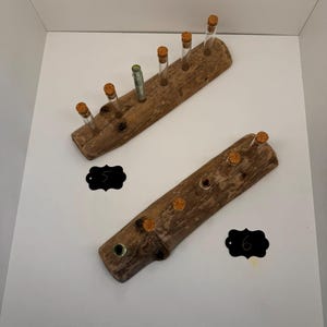 Könnte beinhalten: Zwei Stücke Treibholz mit korkverschlossenen Glasröhrchen, die in gebohrte Löcher eingesetzt sind. Das Treibholz ist braun und hat ein natürliches, rustikales Aussehen. Die Röhrchen sind aus klarem Glas und leer. Das Treibholz ist mit den Zahlen "5" und "6" beschriftet.
