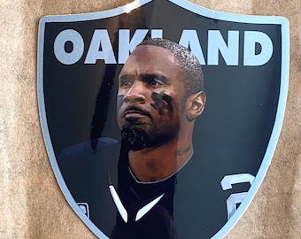 May include: A black and silver shield-shaped sticker with the word "OAKLAND" in silver lettering. A black and white photo of a football player is in the centre of the sticker.