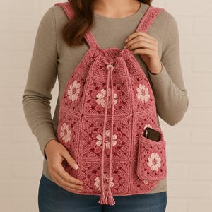May include: A pink crocheted backpack with a drawstring closure and a small pocket. The backpack features a floral pattern in shades of pink and white. The straps are also crocheted. The bag is held by a person.