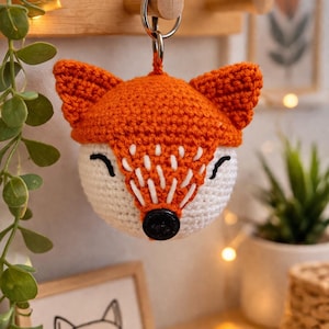 May include: A handmade crochet fox keychain. The fox has an orange head and ears, a white face with black stitched details, and a black button nose. It hangs from a silver keyring. The background includes a wooden peg rack and decorative items.