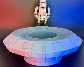 Puede incluir: Un modelo de plataforma circular de plástico gris con un centro elevado y una base circular más pequeña. La plataforma tiene un borde elevado alrededor del borde y una serie de paneles rectangulares en la parte superior e inferior. La plataforma está diseñada para ser utilizada como un expositor para figuras de acción.