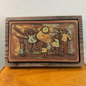 Könnte beinhalten: Eine rechteckige Keramik-Wandplatte mit einem Relief von fünf Kindern, die mit einem Ball spielen, in Braun-, Beige- und gedämpften Grüntönen. Das Kunstwerk ist in verwittertes Holz gefasst und trägt in der Ecke die Initialen PJ.