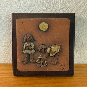 Könnte beinhalten: Eine quadratische Wandplatte aus Terrakotta in einem dunkelbraunen Holzrahmen. Das Relief zeigt zwei Figuren in einer Gartenszene mit einer kreisförmigen Sonne darüber. Eine Figur steht, während die andere sich bückt, um Blumen zu pflegen.