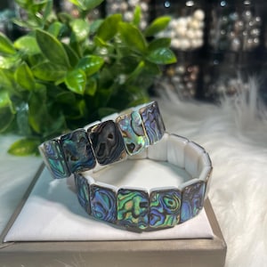 Op de afbeelding: Twee abalone schelp armbanden. Elke armband heeft iriserende, vierkante abalone schelp segmenten. De schelpen tonen een reeks kleuren, waaronder blauw, groen en paars. De armbanden worden gepresenteerd op een wit oppervlak.