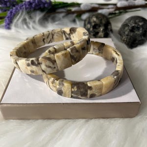 May include: Two square-beaded bracelets. The bracelets are made of light beige and brown speckled stones. The bracelets are resting on a white surface with a beige border. The background includes purple flowers and decorative skulls.