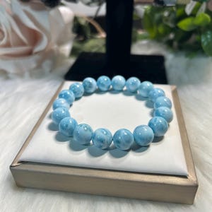May include: A light blue beaded bracelet with spherical beads. The bracelet is displayed on a white surface inside a gold-coloured box. Other bracelets are visible in the background.