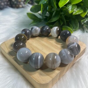 May include: A bracelet made of large, round agate beads, with grey and white stripes. The bracelet is presented on a small, light-coloured wooden block. Green leaves and a white, fluffy surface form the background.