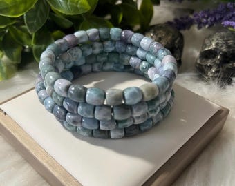 Puede incluir: Tres pulseras apiladas hechas de cuentas azul claro y gris. Las pulseras se exhiben sobre una superficie blanca con un borde marrón. En el fondo se ven follaje verde y flores moradas.